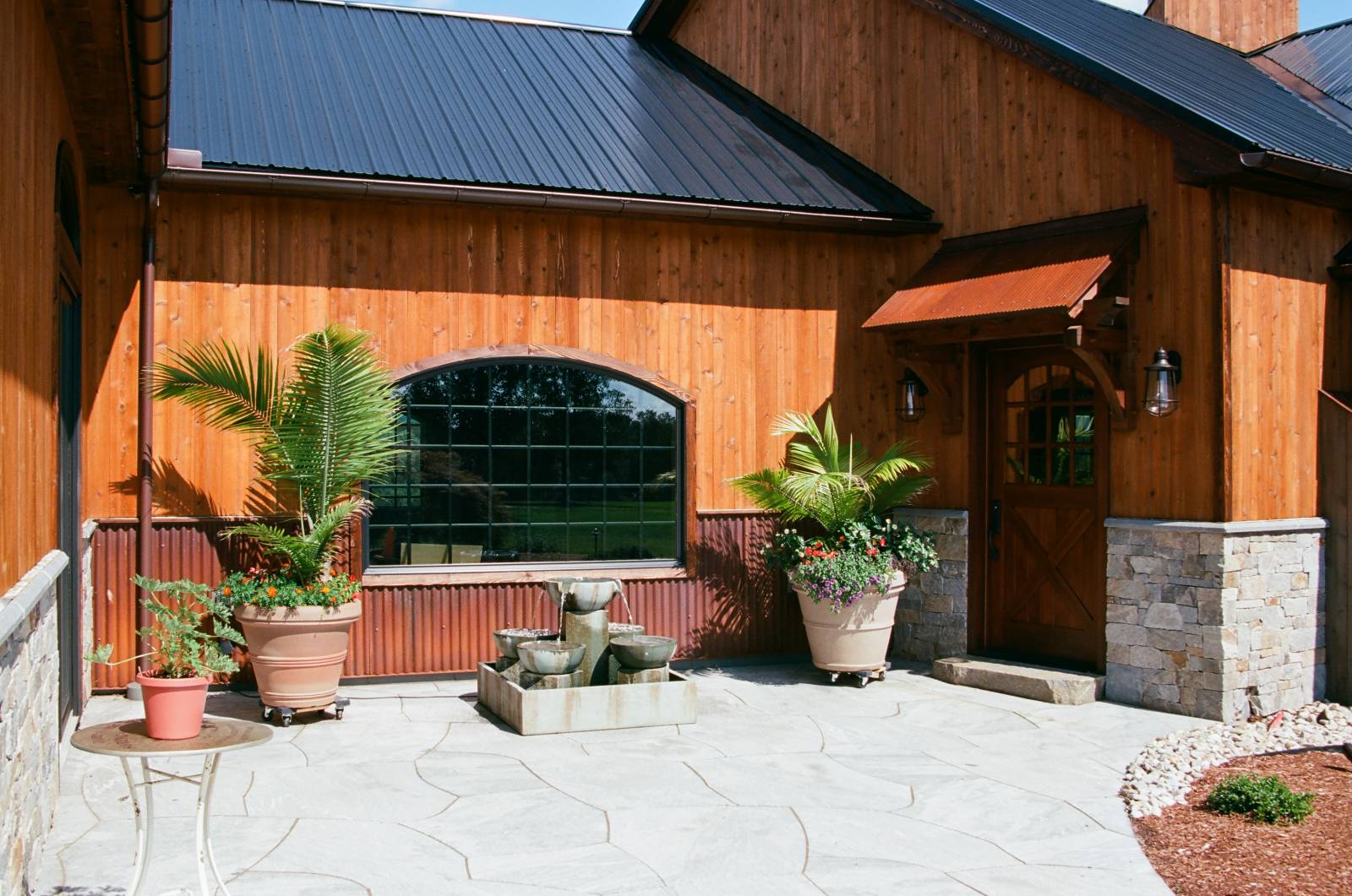 Timber Frame Eyebrow Roof over Entry Door • Arched Bow Top Window • Rustic Siding • Rusty Metal Wainscot • Cultured Stone