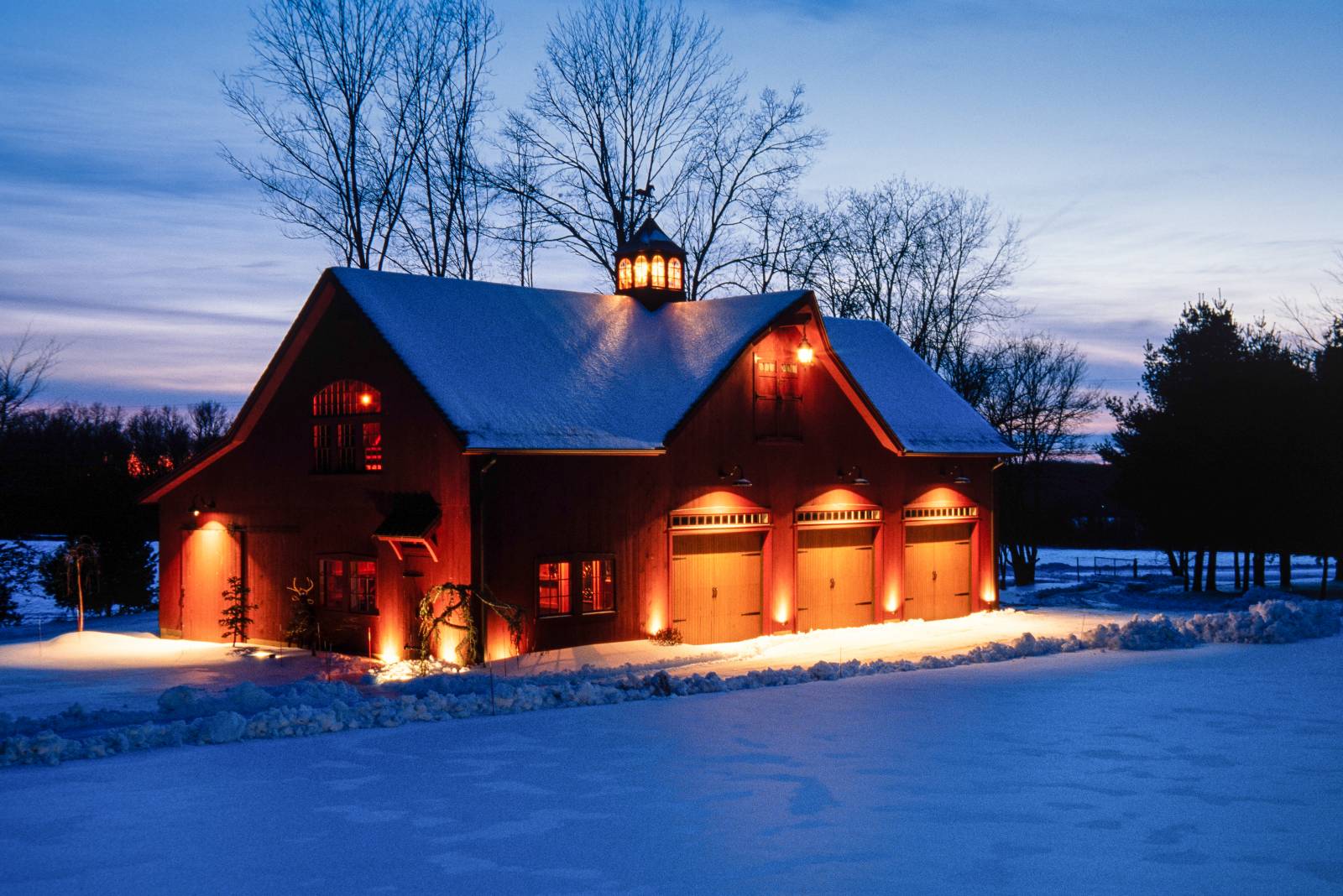 The post & beam Carriage Barn blanketed in snow welcomes with a warm glow after sunset in this peaceful wintry scene in the countryside of Ellington CT